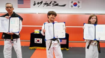 An adult and two young students hold official Kukkiwon Dan and Poom certificates at a black belt ceremony at American Black Belt Academy in Peachtree City, Georgia