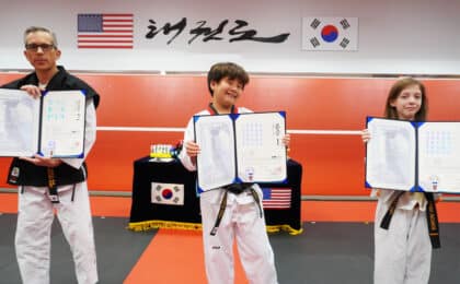 An adult and two young students hold official Kukkiwon Dan and Poom certificates at a black belt ceremony at American Black Belt Academy in Peachtree City, Georgia